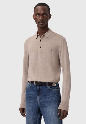 Beżowa koszulka polo z długim rękawem, kołnierzykiem, trzema czarnymi guzikami i małym logo na lewej piersi, zestawiona z niebieskimi jeansami i czarnym paskiem.