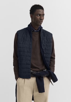 Man wearing navy quilted vest over brown sweater with a scarf tied around waist and beige pants, looking to the side on a plain background.