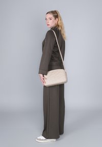 Beige quiltet crossbody taske med en glat tekstur, båret af en person i et mørkt outfit med løse bukser og hvide sneakers.