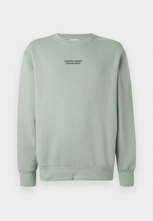 Vaalean salviavihreä crewneck-huppari, jossa on pitkät hihat ja musta teksti "Crafted Goods - Pier One Denim" keskellä rintaa.