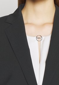 Stříbrná kruhová brož na bílém blůze, upevněná černým blejzrem. Brož má vodorovnou tyčinku uprostřed.