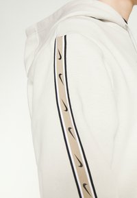 Ljusbeige hoodie med svarta Nike swoosh-logotyper mönstrade på en tanränder längs ärmen, bärs av en person som står vänd åt sidan.