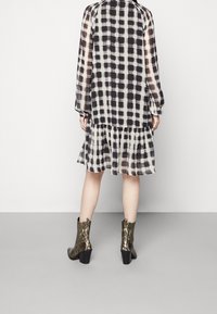 Robe à carreaux noire et blanche avec des manches longues, un ourlet en couches, fabriquée dans un matériau transparent. Portée avec des bottines en cuir serpent métallisé.