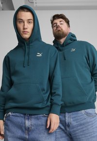 Felpe in cotone color teal di Puma con tasca a marsupio, polsini a coste e ricamo del logo sul petto indossate da due modelli.