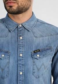 Chemise en denim bleu clair avec un col pointu, des boutons-pression sur le devant, deux poches poitrine avec des détails de couture et une étiquette noire sur la poche.