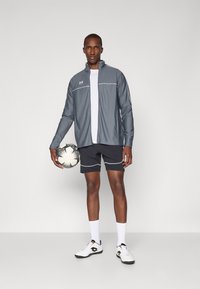 Grey zip-up jacket with white stripes, white t-shirt, black shorts with accents, holding a black-and-white soccer ball, wearing white sneakers.