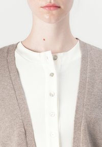 Lichtbruine cardigan over een witte geribde shirt met een ronde halslijn en vijf knopen. Heeft een zachte textuur en een eenvoudig ontwerp.