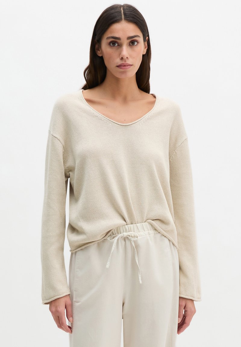 Femme portant un pull en maille beige ample avec un col large et un pantalon beige clair à cordon, debout devant un fond blanc uni.