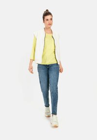 Weißer ärmelloser Reißverschlussweste über einem gelben Langarmshirt, kombiniert mit blauen Jeans und weißen Sneakers. Einfaches Design mit minimalen Akzenten.