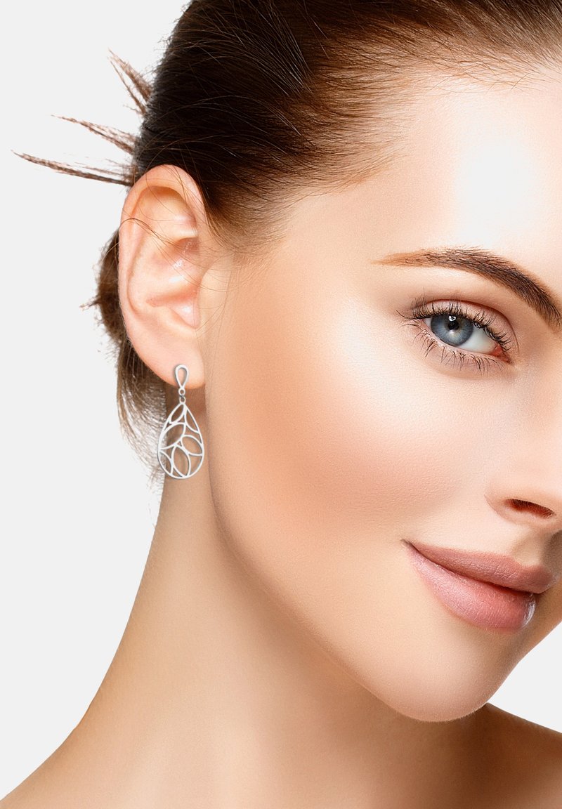 Silverfärgade droppörhängen med bladdesign och intrikata utskärningar, hängande från en ögla, bärs i en modells öra. Slät hudtextur.