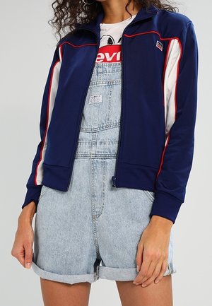 Person iført lyseblå denim-oprullede overaller, hvid grafisk T-shirt og navyblå lynlåsjakke med røde og hvide striber.
