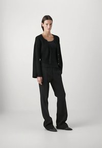 Svart ribbad kofta med knappdetaljer, bärs över en svart linne och kombineras med svarta vida byxor och spetsiga skor.