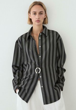 Młoda osoba nosząca luźną czarno-szarą koszulę w pionowe paski i białe spodnie, z rękami w kieszeniach i srebrnym naszyjnikiem z zawieszką.