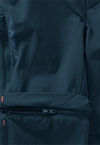 Détail de la veste Vaude bleu marine, avec des poches zippées, un matériau lisse et léger, et des accents orange près des zippers.