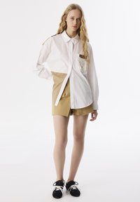 Weißes und beige color-blocked Hemd mit Knopfleiste und Brusttasche, kombiniert mit beigen Shorts und schwarzen Sneakers, vor einem weißen Hintergrund.