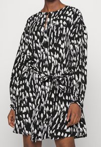 Schwarz-weißes Kleid mit langen Ärmeln, tailliertem Bund und Schlitzkragen. Hat ein abstraktes, malerisches Muster, das das gesamte Kleid ziert.