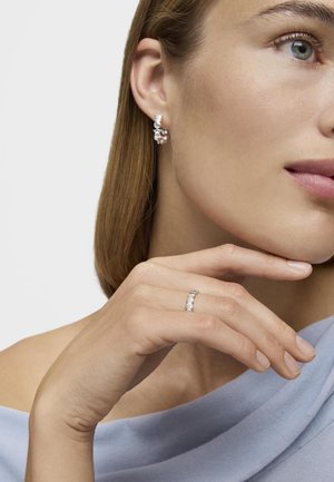 Silver earrings with circular and teardrop shapes, featuring embellishments. A silver ring with small white stones is worn on the hand.