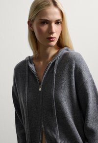 Felpa grigia con zip, caratterizzata da un tessuto morbido e testurizzato, un cappuccio e polsini a costine. Il capo presenta un design rilassato con dettagli di cucitura sottili.