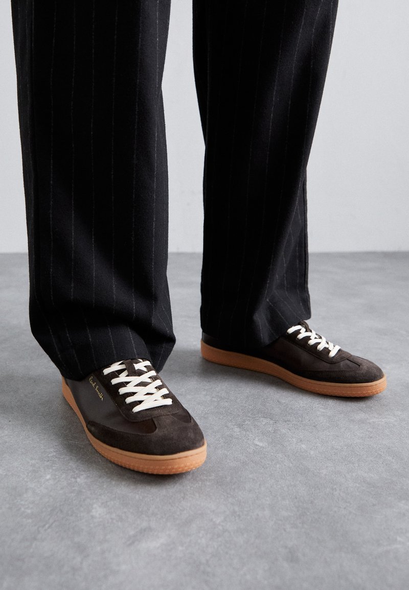 Baskets en suède marron foncé avec des lacets crème, des accents blancs et une semelle en caoutchouc beige. Portées avec un pantalon noir à rayures.