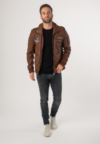 Braune Lederjacke mit Reißverschluss, schrägem Kragen und mehreren Patches auf der Brust und dem Arm; kombiniert mit schwarzem Shirt, grauen Jeans und weißen Sneakers.