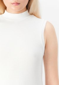 Detailní záběr na osobu se světlou pletí a blond vlasy, která má na sobě bezrukávový bílý top s vysokým límečkem na bílém pozadí.