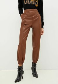 Pantaloni in pelle sintetica marrone con vita alta e polsini elasticizzati. Abbinati a stivali neri con punta a stabetto e dettagli in catena.