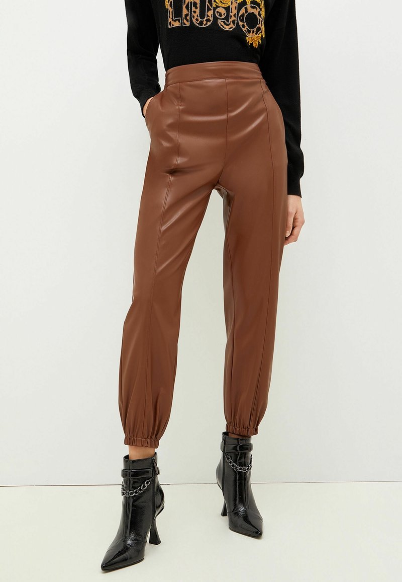 Pantaloni in pelle sintetica marrone con vita alta e polsini elasticizzati. Abbinati a stivali neri con punta a stabetto e dettagli in catena.