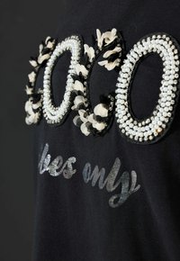Schwarzes T-Shirt mit dem Schriftzug "Coco" in verzierten Texturen mit Pailletten, Blumenakzenten und einem metallischen Glanz mit der Aufschrift "loves only".