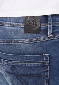 Jeans in denim blu con una toppa in pelle con un design in rilievo. Il tessuto ha una texture consumata e cuciture visibili lungo le cuciture.