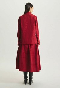 Tenue rouge avec une chemise à boutons et une longue jupe superposée. Le tissu semble lisse avec une finition mate. Des bottes noires jusqu'aux genoux complètent le look.