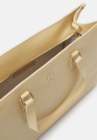 Sac fourre-tout beige clair avec une texture en cuir lisse, doté d'une fermeture éclair, de quincaillerie dorée et de poignées doubles pour le transport. L'intérieur est doublé.