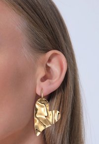 Guld hjerteformet ørestik med en struktureret, bølget overflade, fastgjort med en lille ring, præsenteret mod en lys baggrund.