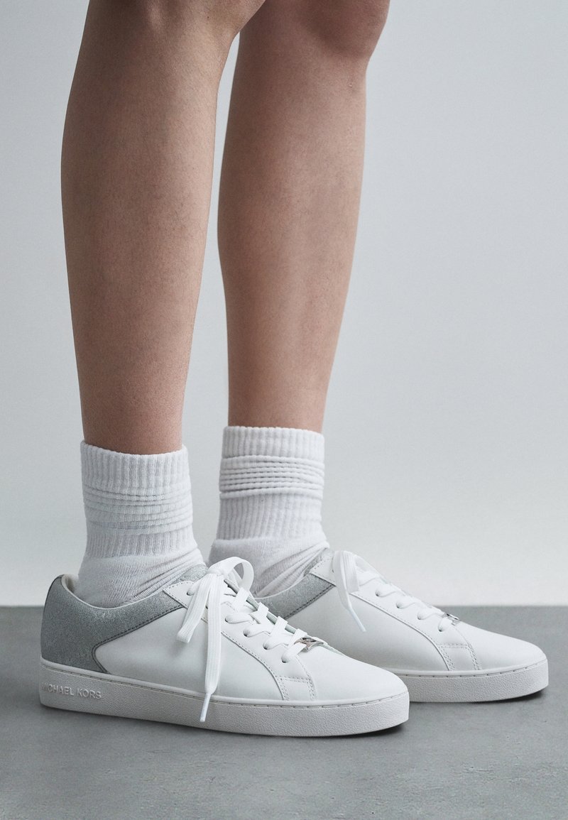 Personne portant des baskets blanches avec des accents gris au talon et des chaussettes blanches côtelées, debout sur un sol gris sur fond uni.