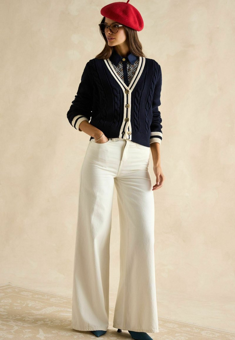 Jūras spēku krāsas adīts kardiganis ar baltajām akcentiem, krēmkrāsas platās kājas bikses un sarkans berets. Nēsātāja stāv ar roku uz gurniem, izceļot apģērbu.