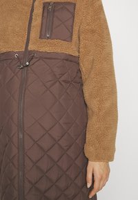 Καφέ καπιτονέ μπουφάν με αμάνικο τμήμα σε μπεζ χρώμα. Διαθέτει μια τσέπη στο στήθος με φερμουάρ και ρυθμιζόμενο κορδόνι στη μέση.