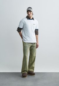 Bela majica s modrimi poudarki, čez njo na plastih karirana majica z dolgimi rokavi, kombinirana z zelenimi hlačami s črtami in rjavimi čevlji. Sivi beanie na glavi.