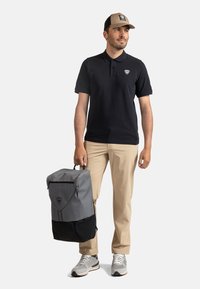 Černé polo tričko, béžová kšiltovka, světlé kalhoty, šedý a černý batoh se zipem a šedé tenisky. Batoh má logo a dvoubarevné sekce.