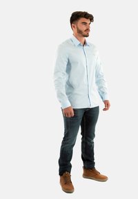 Camisa de botones de manga larga azul claro con una textura suave, combinada con jeans oscuros y botas marrones. La camisa tiene un cuello en contraste.