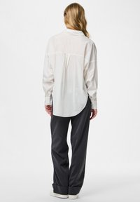 Camisa blanca de manga larga con dobladillo redondeado y detalle en el yugo trasero, combinada con pantalones gris oscuro de pierna ancha y zapatillas blancas.