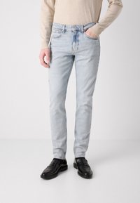 Világoskék, slim fit farmernadrág, elhasználódott textúrával. Klasszikus öt zsebes kialakítással és gombos záródással rendelkezik. Fekete cipővel viselve.