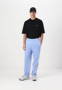 Black t-shirt with "ALLSAINTS" logo, light blue sweatpants with embroidered "Colucci," elastic waistband, and gray sneakers.