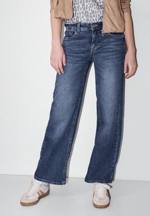 Osoba ubrana w szerokie niebieskie dżinsy, białe trampki z beżowymi akcentami, koszulę w panterkę i beżową kurtkę, stojąca przy białej ścianie.