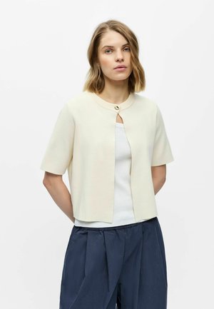 Femme aux cheveux châtain clair porte un cardigan crème à manches courtes boutonné par-dessus un haut blanc et un pantalon bleu marine ample sur un fond blanc uni.