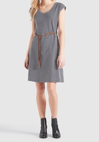 Femme portant une robe rayée verticalement noire et blanche avec un col en V, une ceinture tressée marron et des bottines noires, debout sur un fond uni.