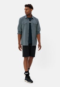 Maglietta grigia leggera a maniche lunghe con due tasche sul petto, indossata sopra una T-shirt blu scuro. Pantaloni corti neri e scarpe da ginnastica completano l'outfit.