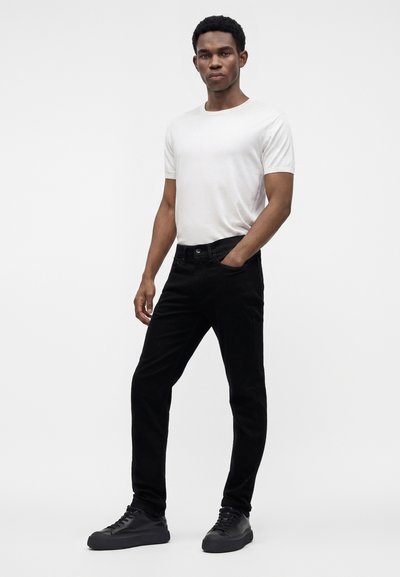 Homme debout, une main dans la poche, portant une chemise blanche à manches courtes, un pantalon noir et des baskets noires sur un fond clair uni.