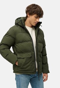 Giacca a vento verde con cappuccio, dotata di chiusura a zip, tasche frontali e texture trapuntata. Indossata sopra un maglione di colore chiaro.
