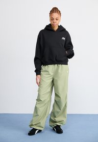 Black hooded sweatshirt with a front pocket, paired with loose, light green trousers and black athletic shoes on a blue surface.