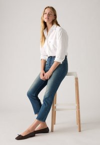 Balta marškiniai su mygtukais, mėlynos aukštos liemens džinsai su išblukusia danga ir rudos plokščios bateliai. Sėdi ant medinio suolo prieš neutralų fono.