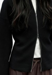 Veste noire en maille avec une fermeture éclair argentée, présentant une coupe ajustée et des manches légèrement évasées, portée sur un haut blanc.